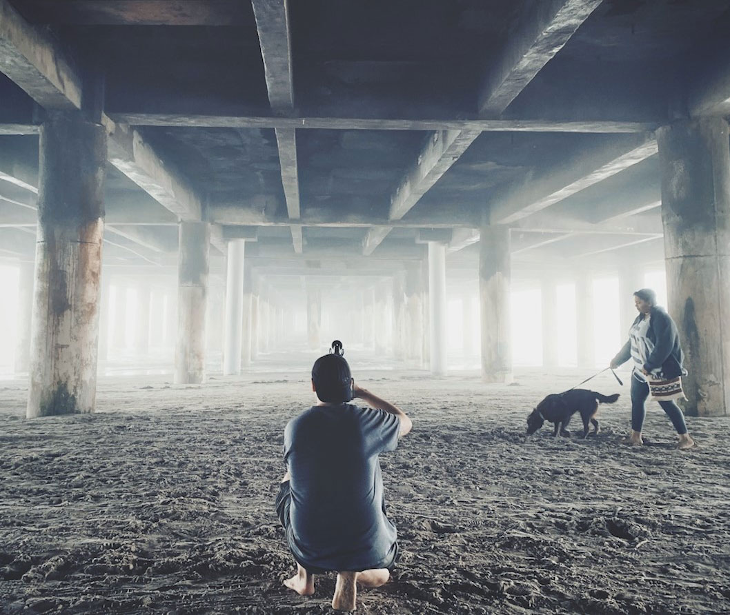 A photographer kneels down in the sand while taking a photo of a woman walking her dog beneath a pier by the ocean.