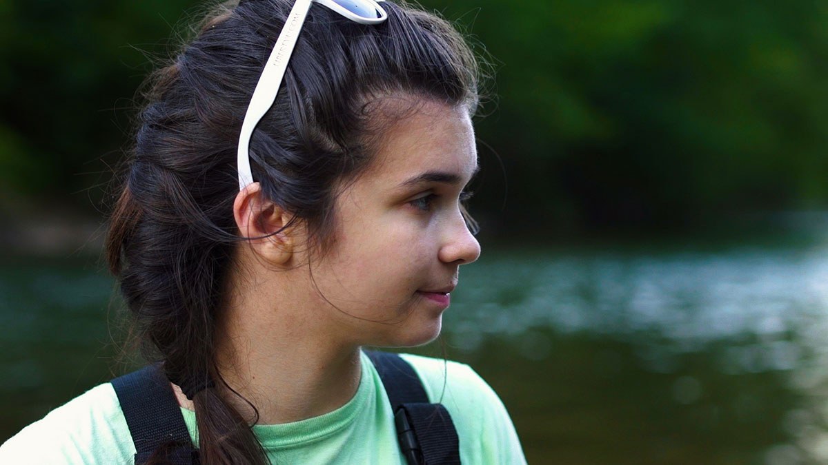 Close up of Megan Bolich in a green tshirt with white-framed sunglasses 					on her head in front of stream where she was flyfishing with her father.