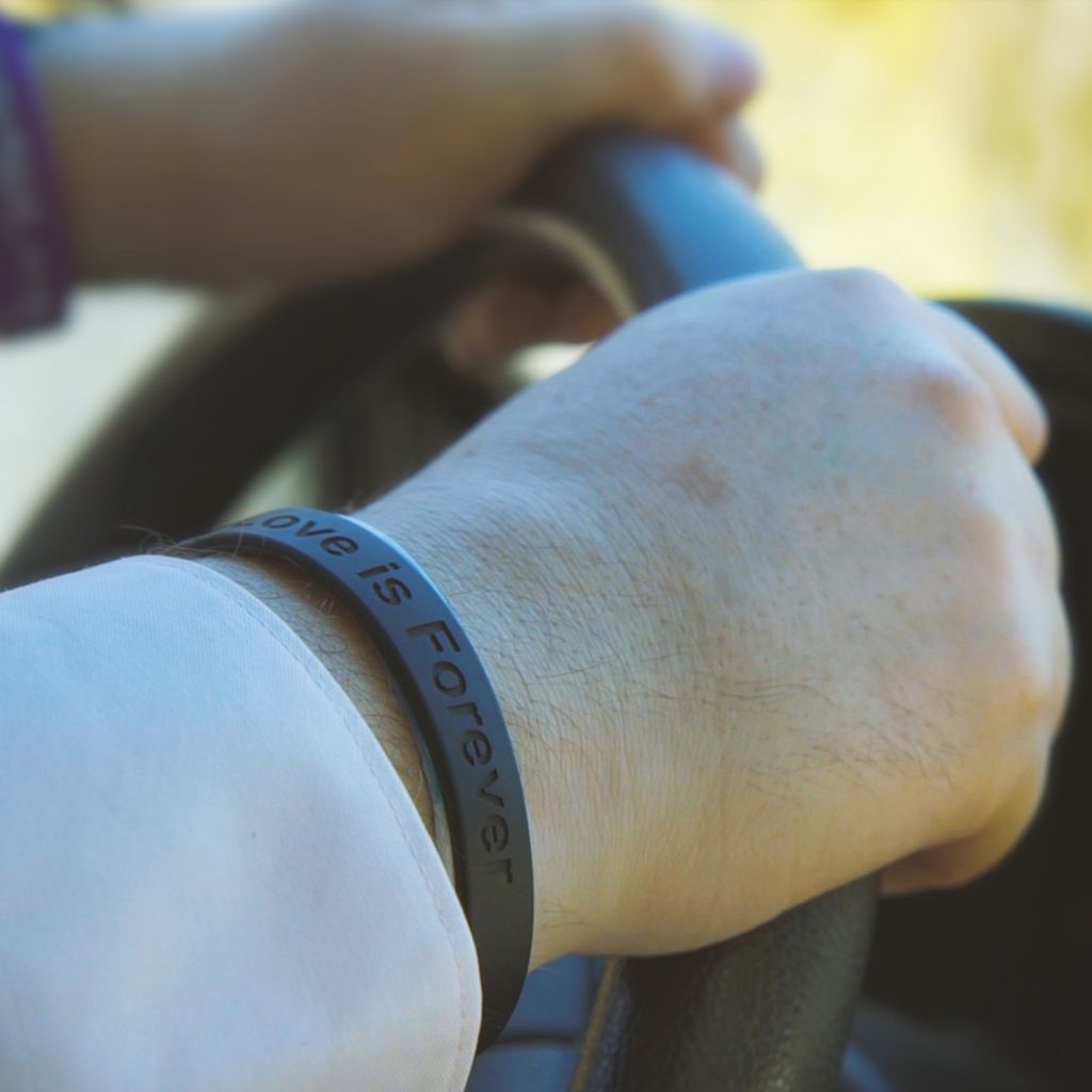 Closeup of a pair of hands on a steering wheel. On the right wrist is a black bracelet inscribed with the words, Love is Forever